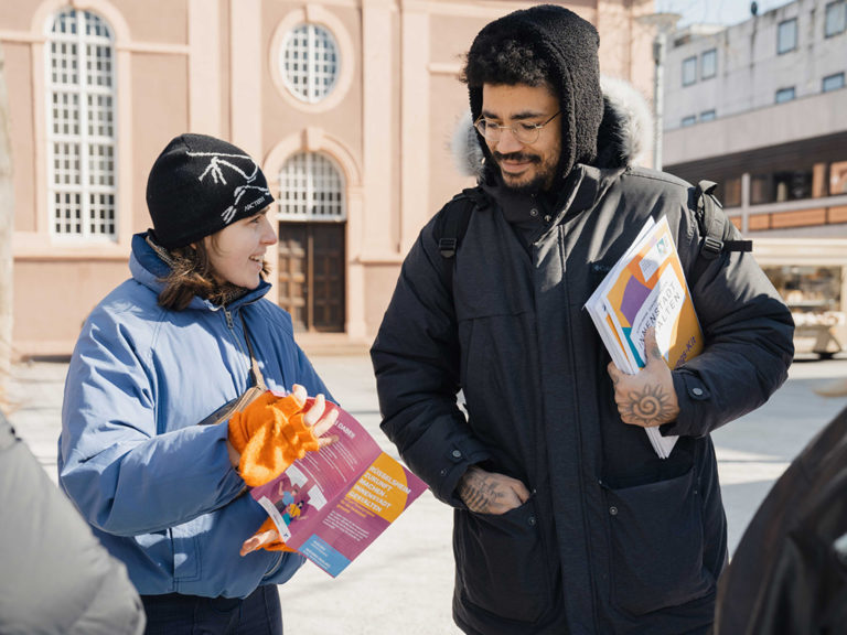 Eine Mitarbeiterin des Projektteams spricht mit einem Passanten und erklärt anhand des Informationsmaterials den Prozess